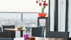 A restaurant with a high table and chairs, a menu stand, a fan, and a potted plant with red flowers. - Continent Centrepoint Hotel Makassar (Makassar)