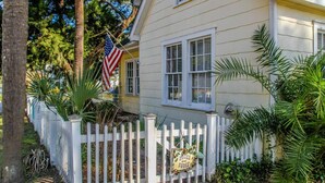 Una casa gialla con una recinzione bianca a doghe e una bandiera americana, con un cartello che dice 'Roses for Sale'.
