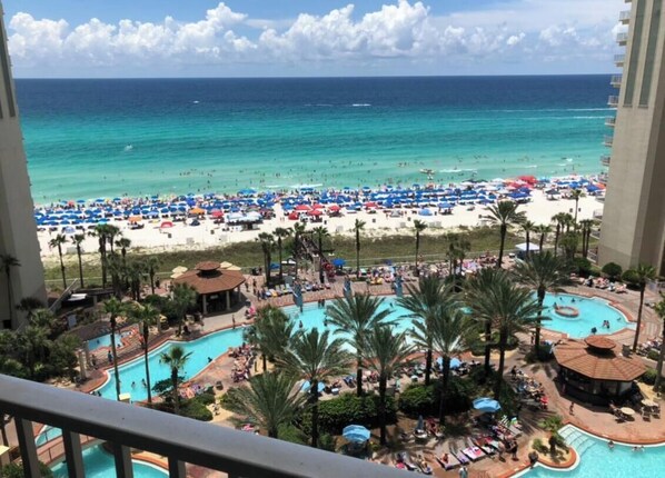 A view from a balcony overlooking a beach with a pool area, palm trees, and beach umbrellas.
