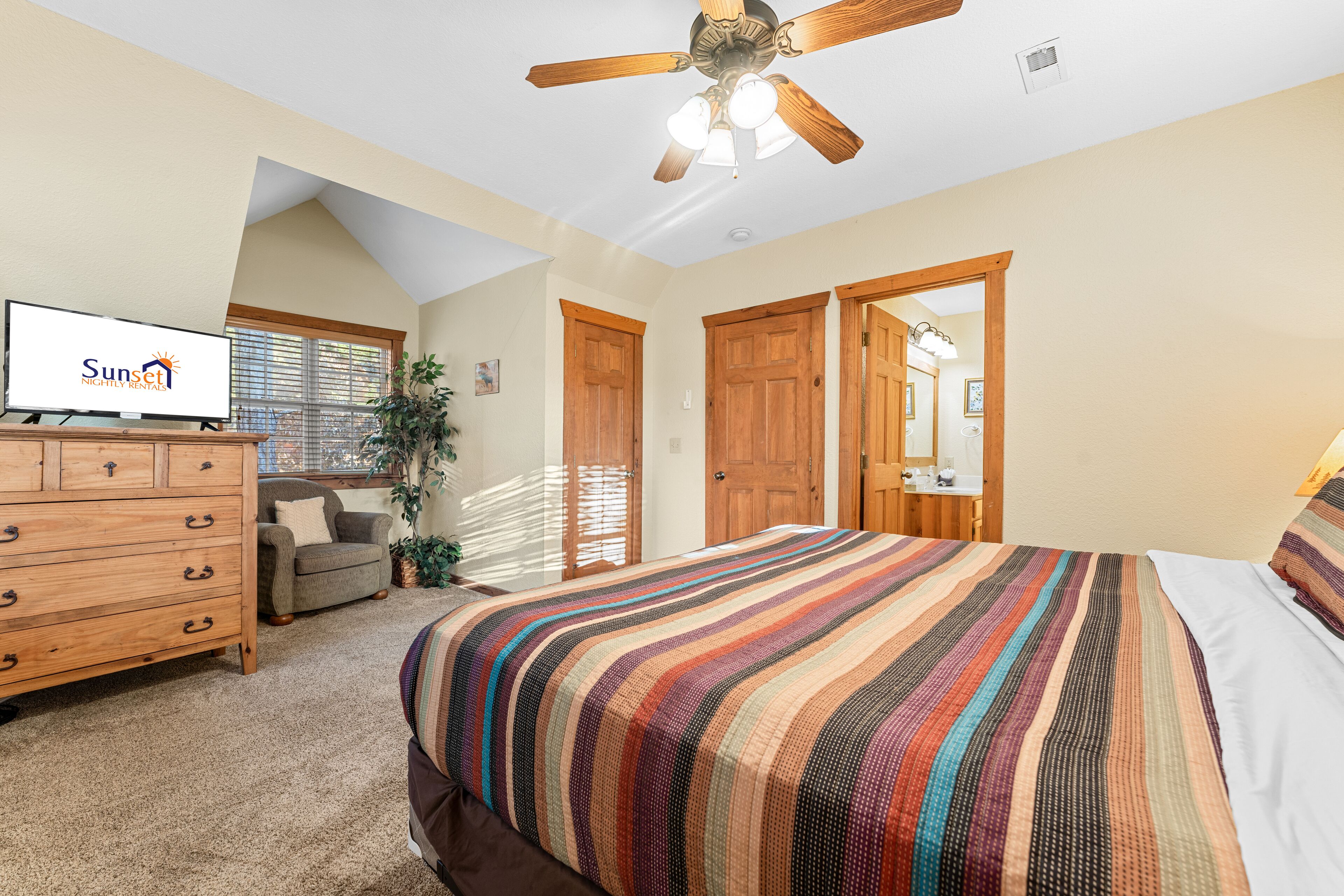 A bedroom with a bed, a dresser, a ceiling fan, and a television displaying "Sunset Nightly Rental".