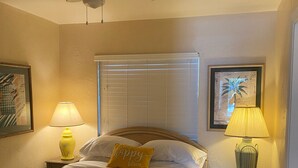 Una habitación de hotel con una cama, dos lámparas de noche, un ventilador de techo, una ventana con persianas y una puerta.