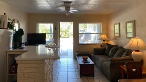 Una sala de estar con sofá, una mesa de centro, televisor y un área de cocina con refrigerador y mostrador.