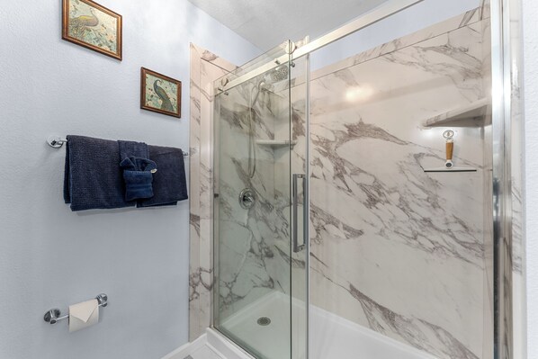 Un baño con ducha envuelta en vidrio, paredes de mármol, un toallero con una toalla doblada y dos cuadros en la pared.