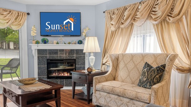 A living room with a fireplace, a sofa, a coffee table, and a television displaying 'Sunset Nightly Rentals'. - Uniquely charming Pet Friendly 2 Bed condo near the strip at Pointe Royale! (Branson)