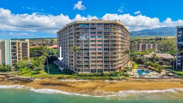 A multi-story hotel with a curved facade, located near a beach with a pool and lounge area.