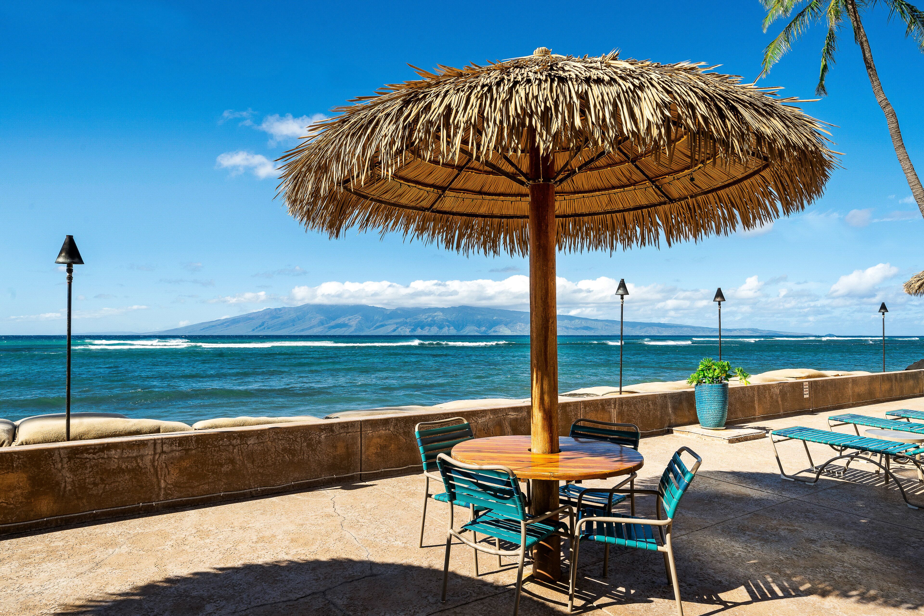 Ein Sitzbereich am Strand mit Tisch und Stühlen, einem Reetdachschirm und Blick auf das Meer und die Berge.
