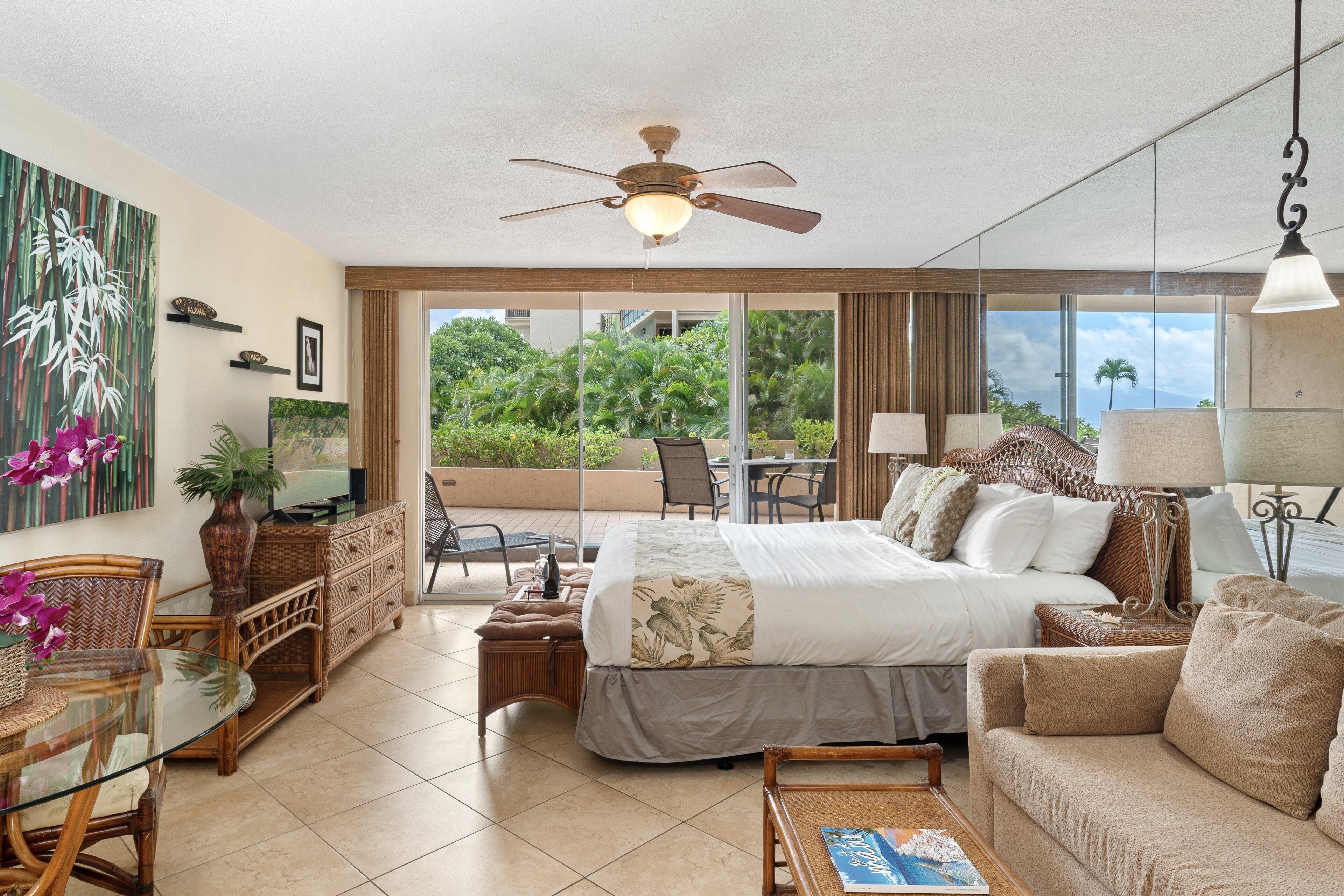 A hotel room with a bed, a desk, a chair, a sofa, a dining table, a ceiling fan, and a view of the outdoors.
