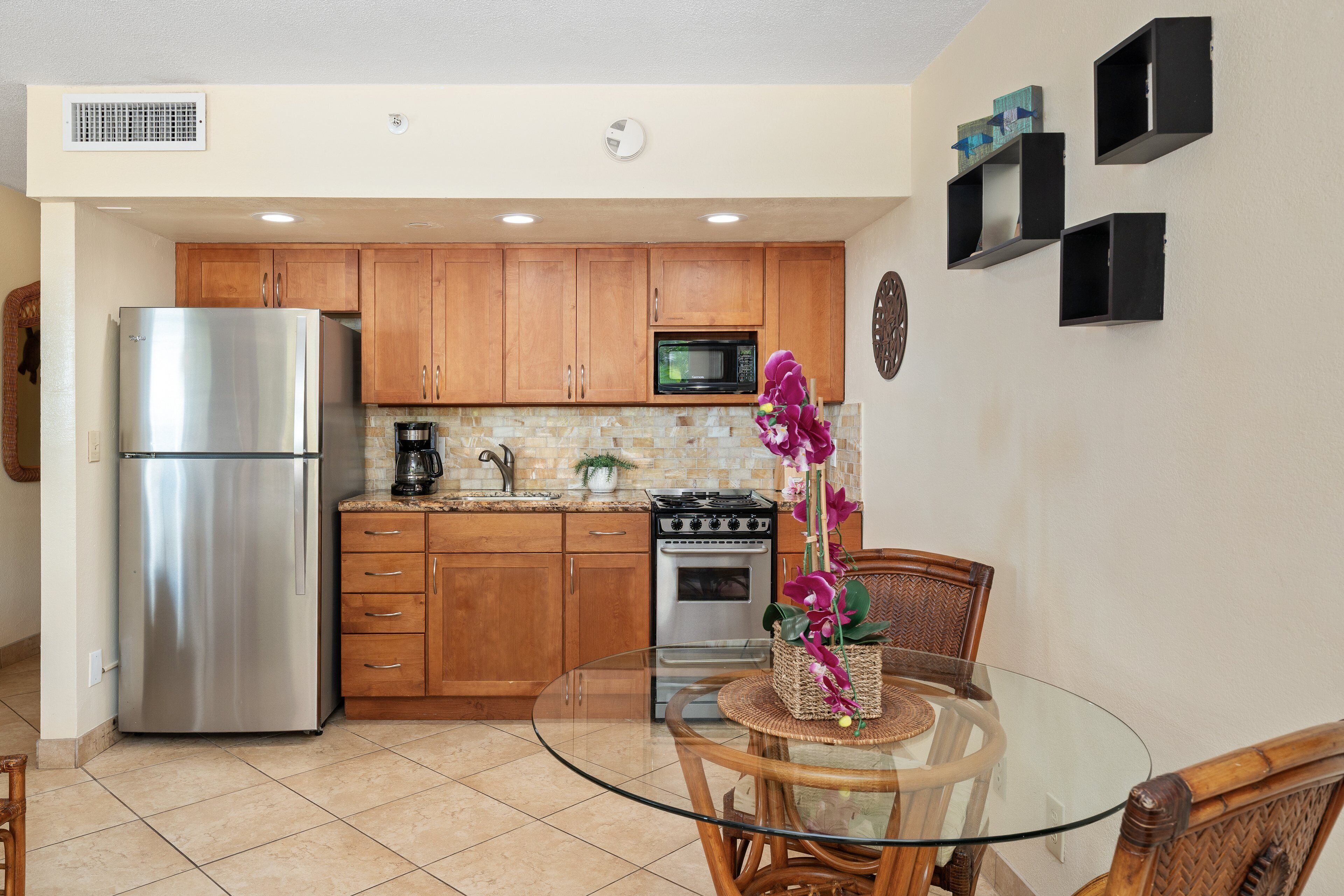 A kitchen with wooden cabinets, a stainless steel refrigerator, a microwave, a stove, and a glass table with a chair.