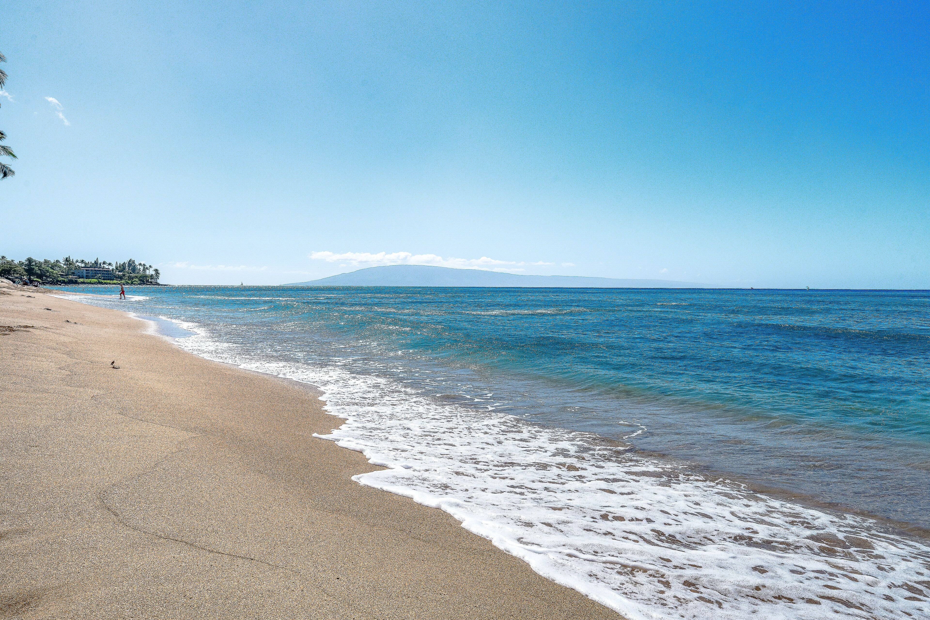 A beach with clear blue water, a sandy shore, and a distant mountain.