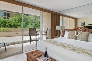 A hotel room with a bed, a small table with a bottle of wine, a chair, and a view of a balcony with plants. - West Maui Studio at BeachFront Resort w Pool- Sleeps 3 Royal Kahana 104 (Lahaina)