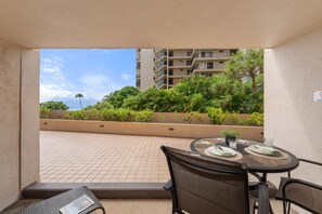 A balcony with a table set for dining, overlooking a landscaped area with trees and a building in the background. - West Maui Studio at BeachFront Resort w Pool-Royal Kahana 104 (Lahaina)