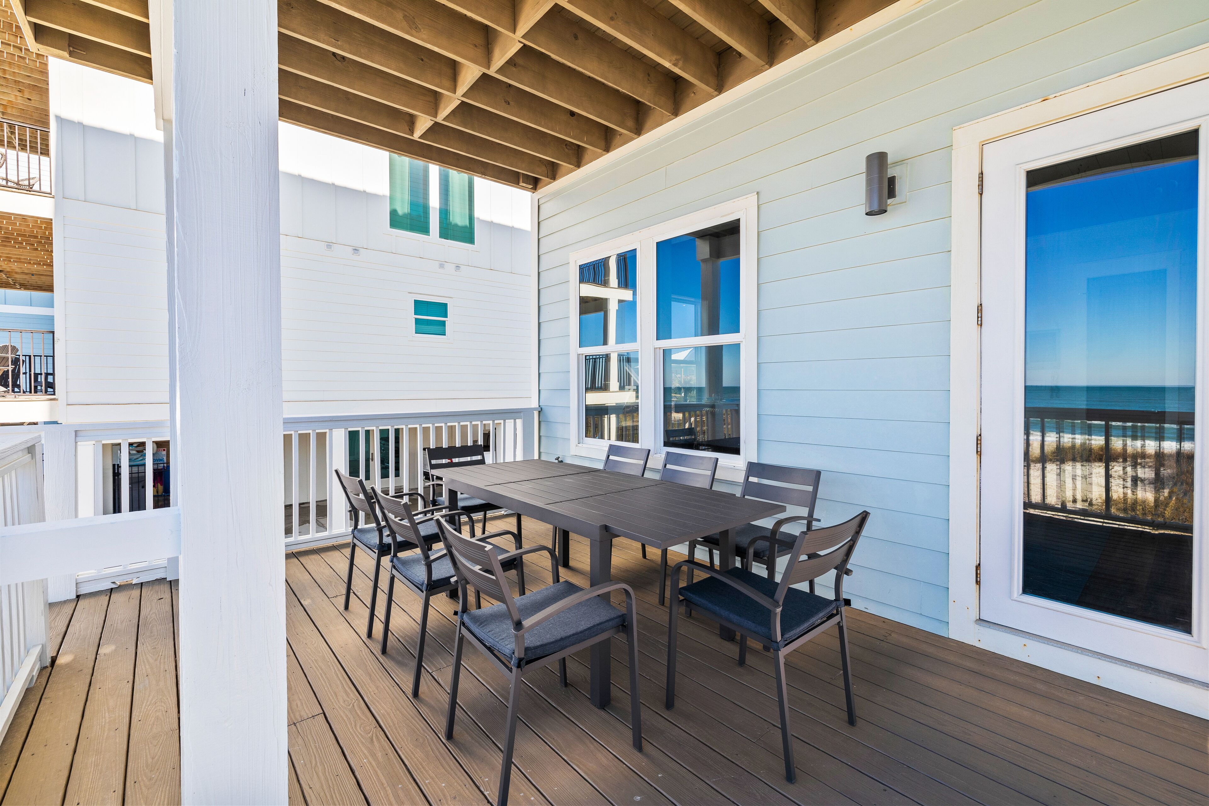 A covered outdoor deck with a table and chairs, a railing, and a view of the beach through a large window.