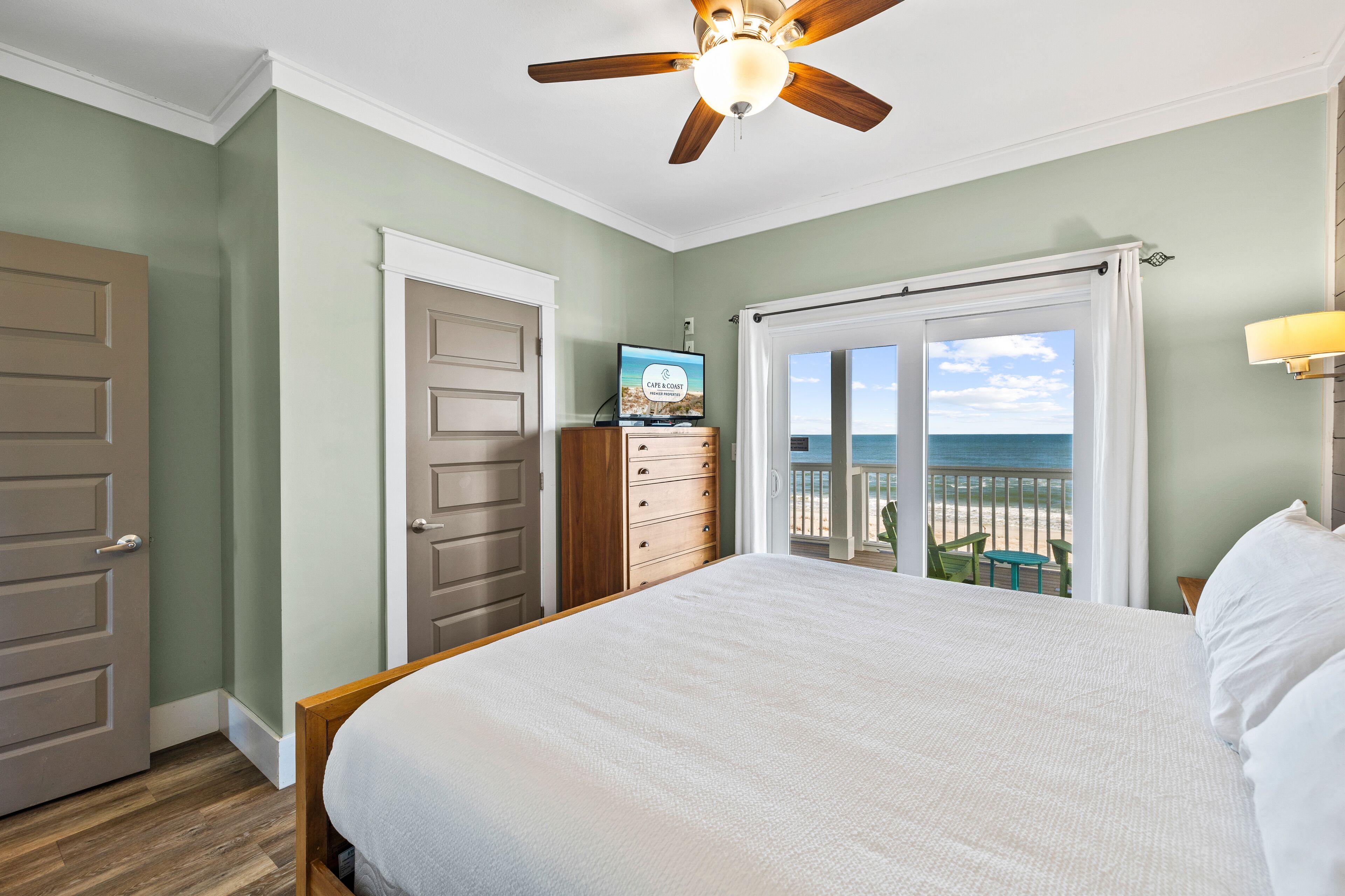 A bedroom with a bed, a dresser, a ceiling fan, a TV, and a view of the beach through a sliding door.