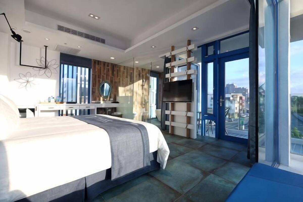 A modern bedroom with a large bed, a desk, and a view of the city through large windows.