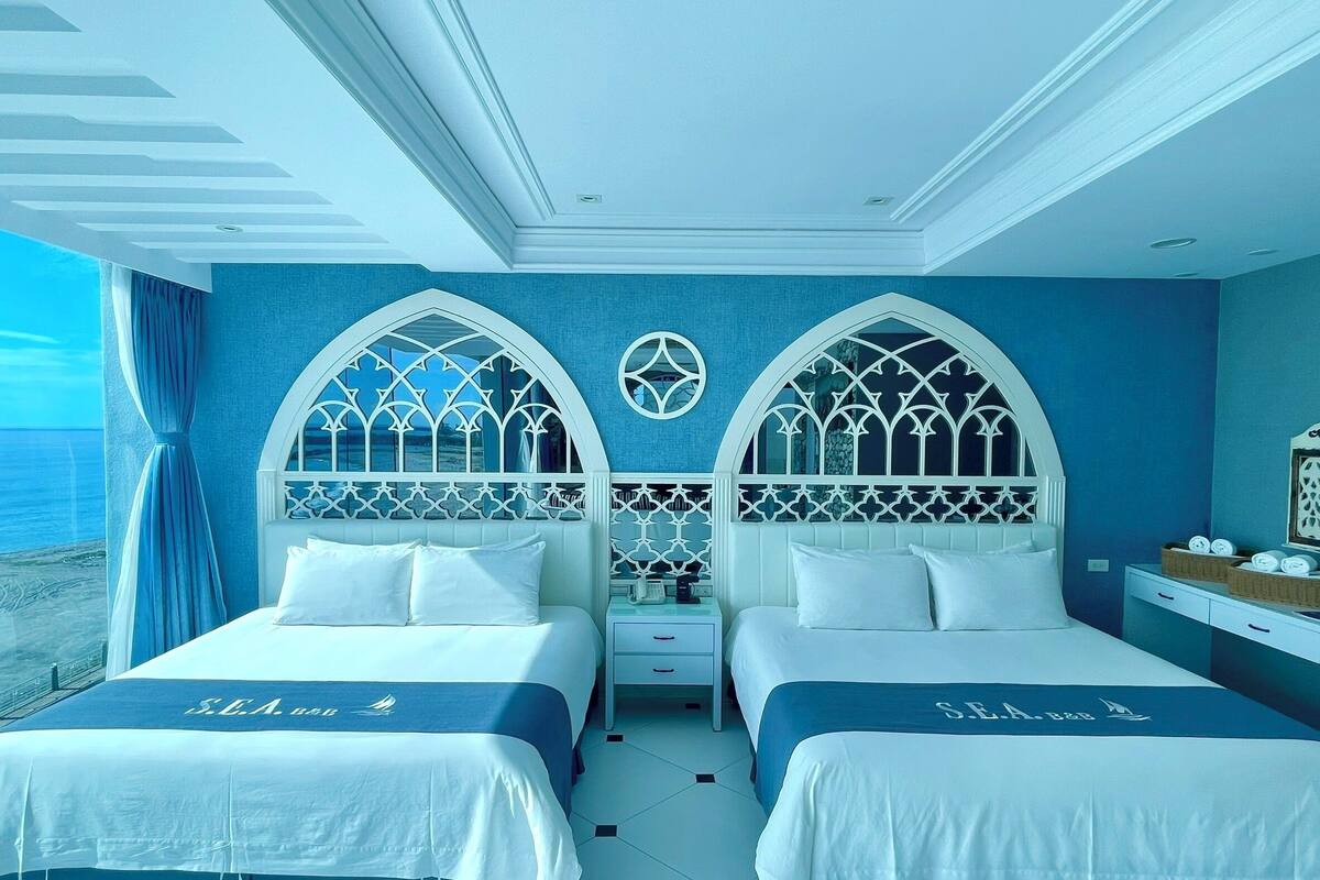 A room with two beds, each with a white headboard and a blue bedspread. There is a small table with a mirror and a basket on it.