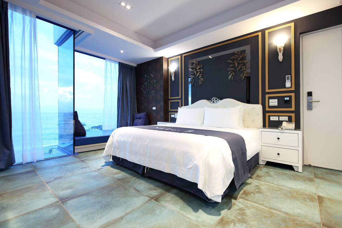 A modern hotel room with a large bed, a nightstand, a wall-mounted lamp, and a view of the ocean through a sliding door.