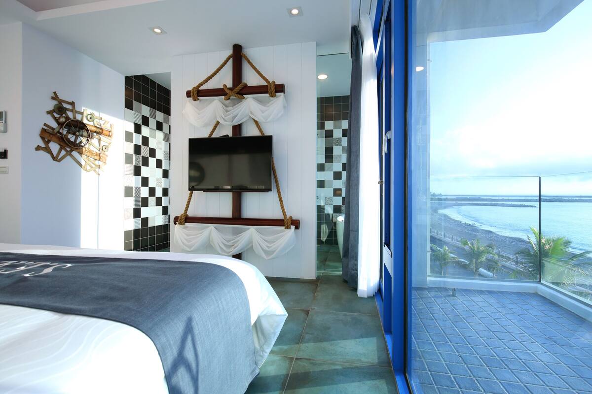 A modern hotel room with a large bed, a TV mounted on the wall, and a view of the beach through a balcony.