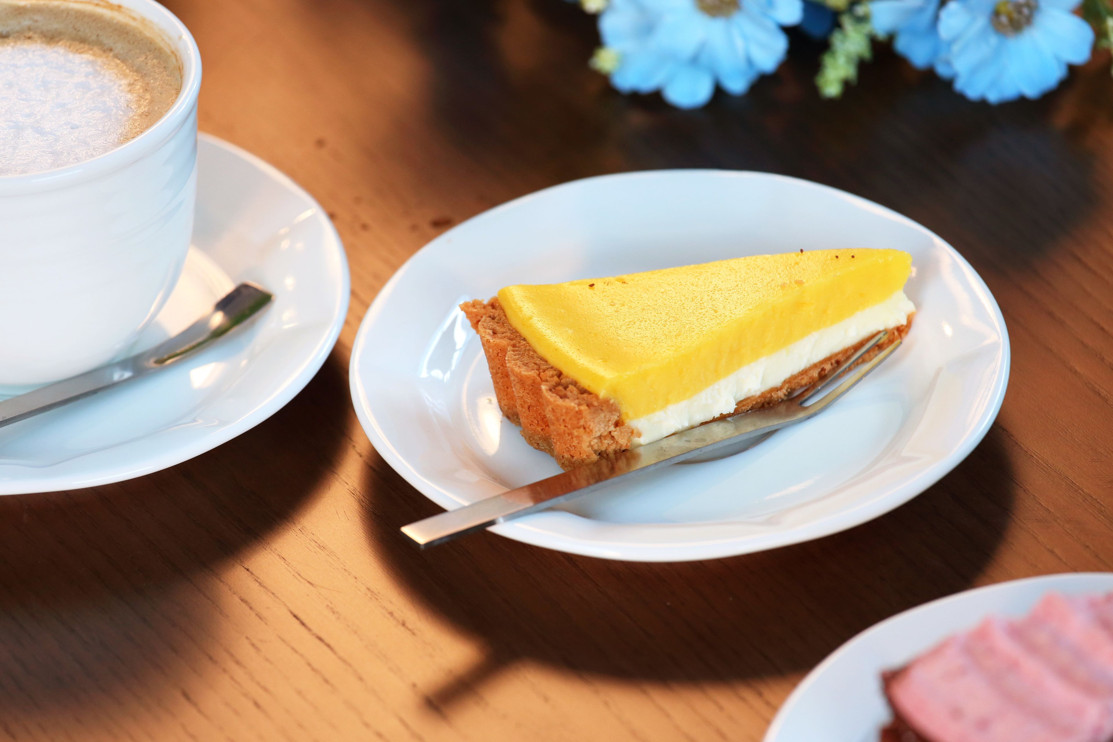 A slice of cheesecake on a white plate with a knife, a cup of coffee, and a plate with ham.