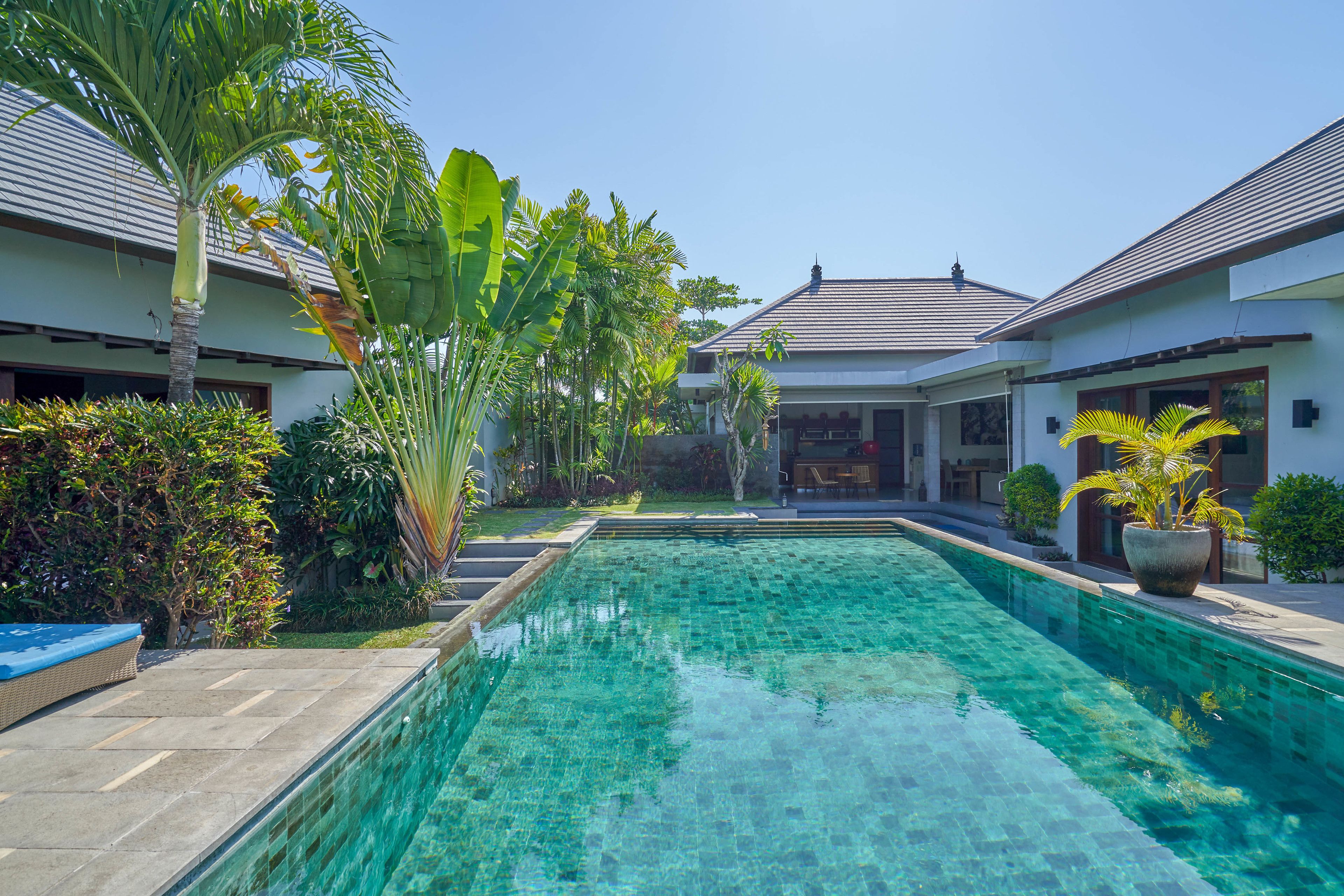 A swimming pool with clear blue water, surrounded by lush greenery and a modern house with a tiled roof.