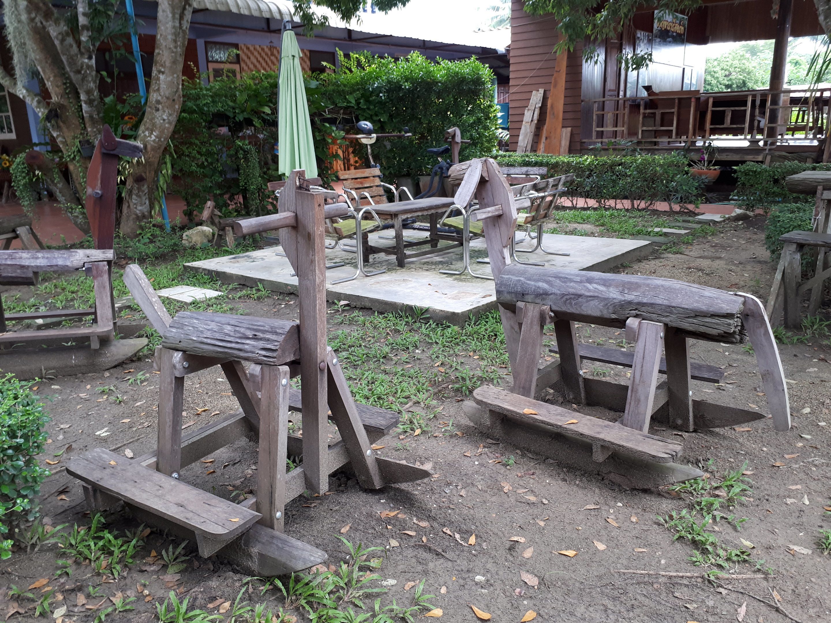 A rustic outdoor seating area with wooden benches and tables, surrounded by greenery and a building in the background.