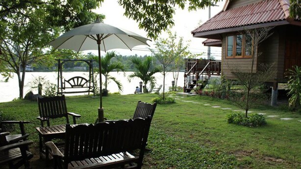 A wooden bench and chairs under an umbrella by a lake, with a house and a small garden in the background. - Chantara Resort (Bang Khonthi)