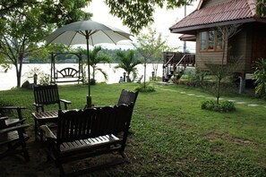 A wooden bench and chairs under an umbrella by a lake, with a house and a small garden in the background. - Chantara Resort (Bang Khonthi)