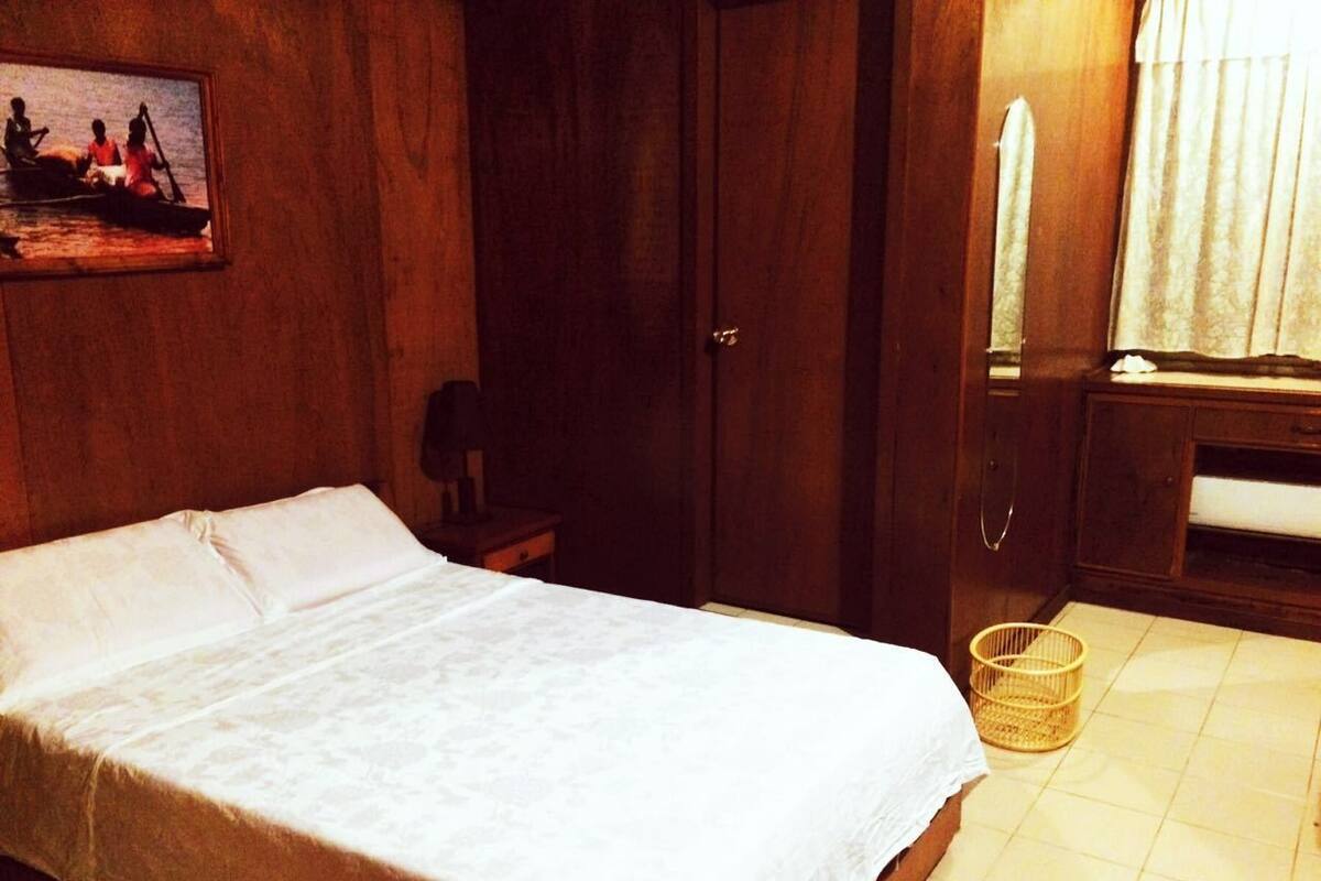 A single bed in a room with wooden walls, a small bedside table, a basket, and a window with curtains.