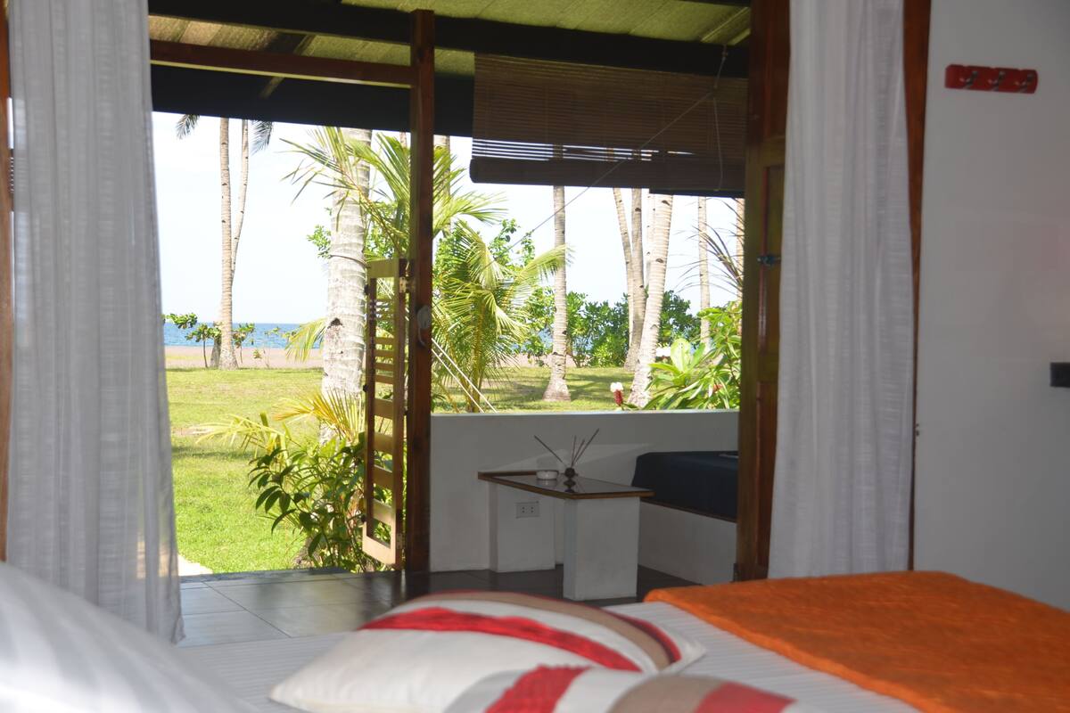 A room with a bed, a small table, and a view of the beach and palm trees.