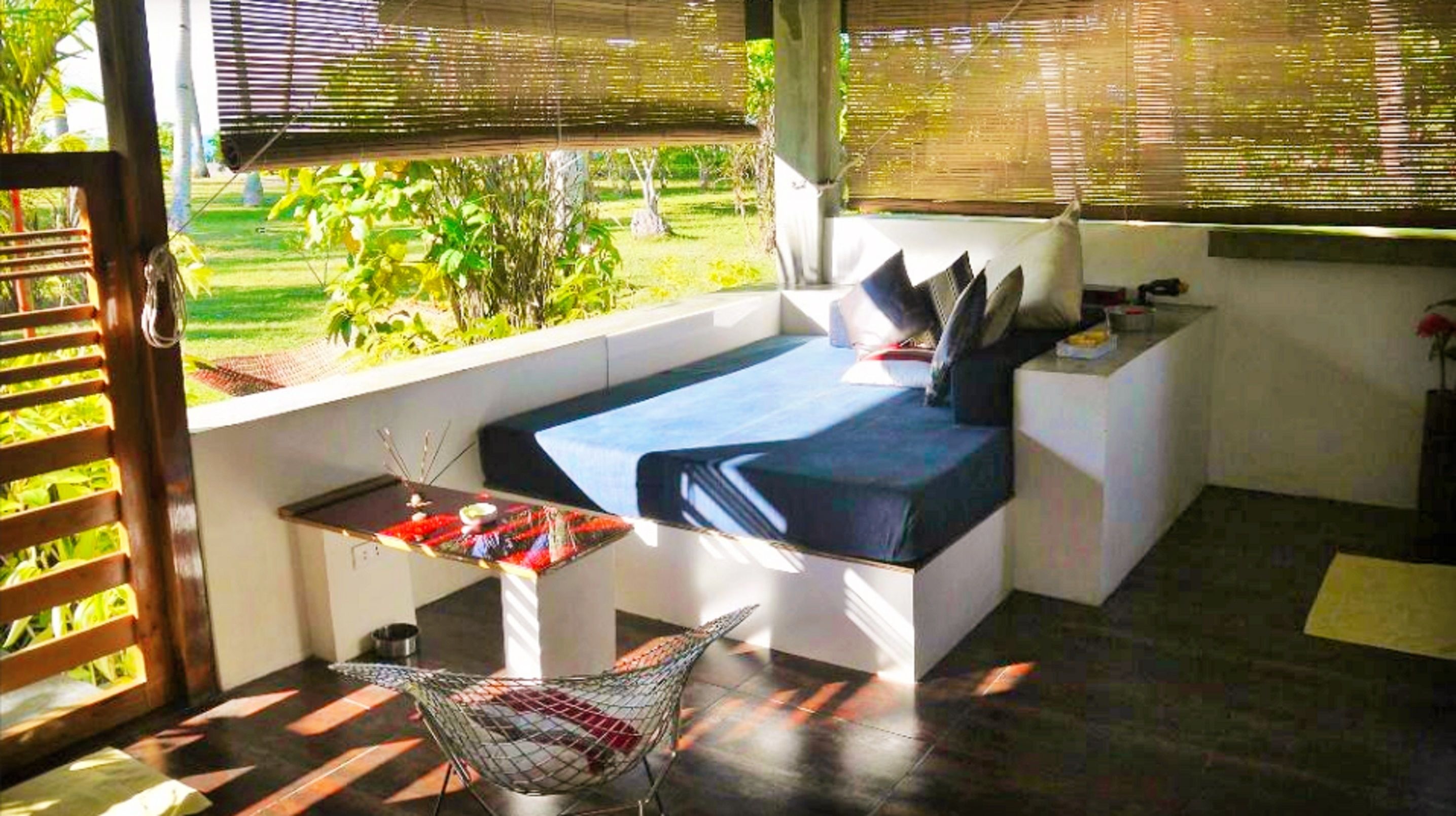 Ein Zimmer mit Bett, einem kleinen Tisch und einer Hängematte. Ein Fenster mit Jalousien bietet Blick ins Grüne.