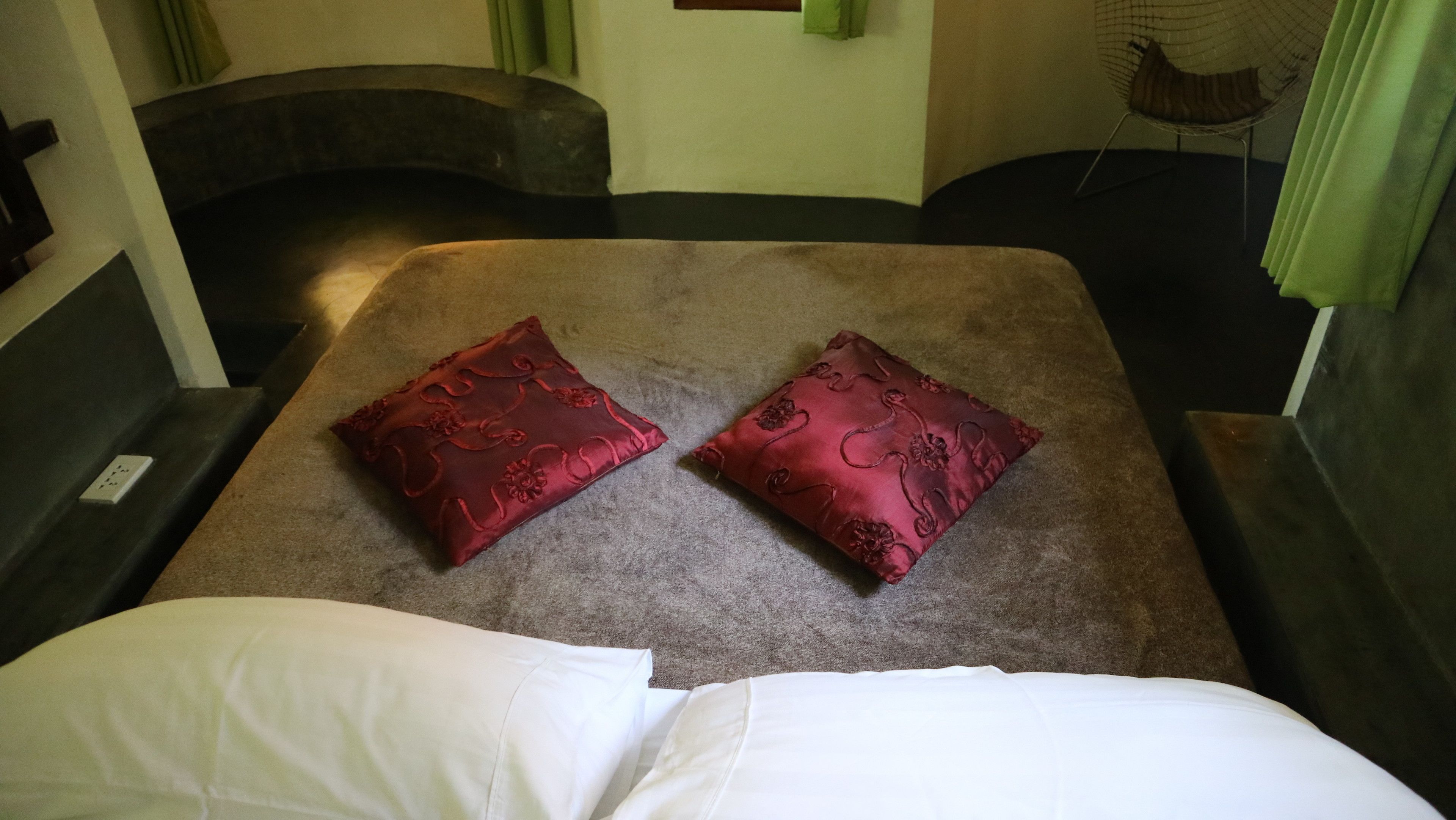 A bed with two red pillows and a white pillow, a small table, and a chair with a green curtain.