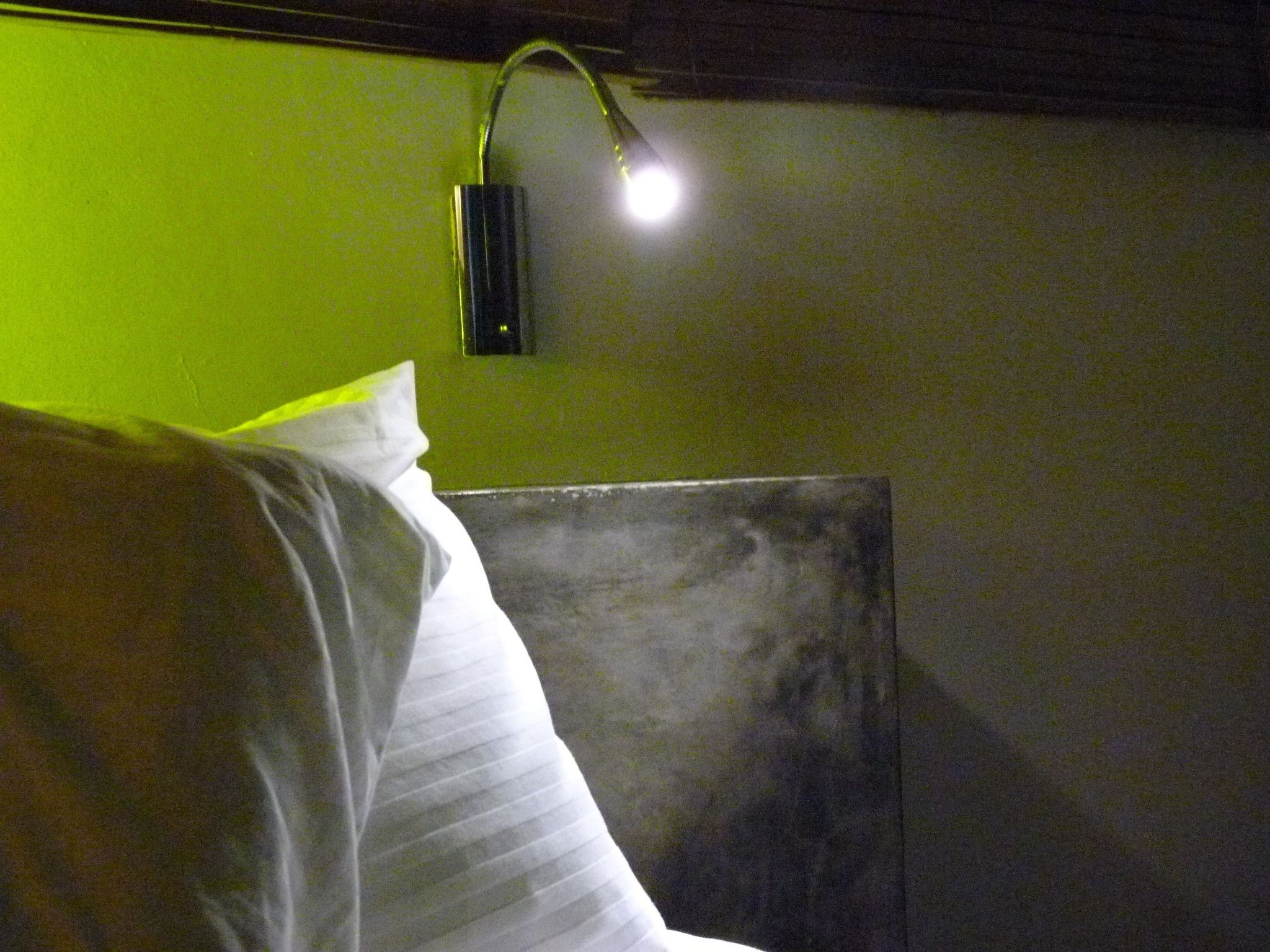 A bed with white bedding and a headboard against a green wall. A wall-mounted light fixture is visible above the bed.