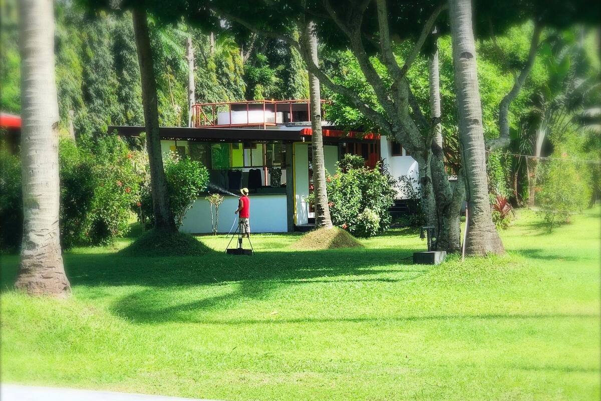 A small, single-story building with a balcony, surrounded by lush greenery and trees.