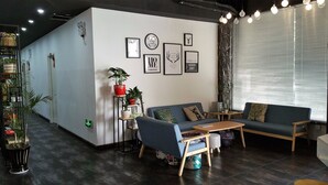 A modern living room with a blue sofa, a wooden coffee table, and a small side table. There are several framed pictures on the wall, a potted plant, and a window with blinds. - Suzhou Chope Hostel (Suzhou)
