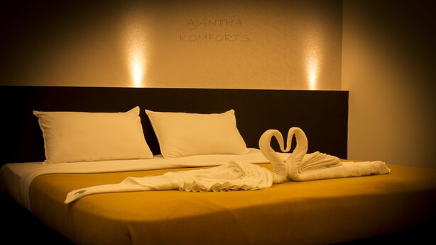 A neatly made bed with white linens and a towel arranged in a heart shape on a yellow bedspread. - Ajantha Komforts (Bengaluru)