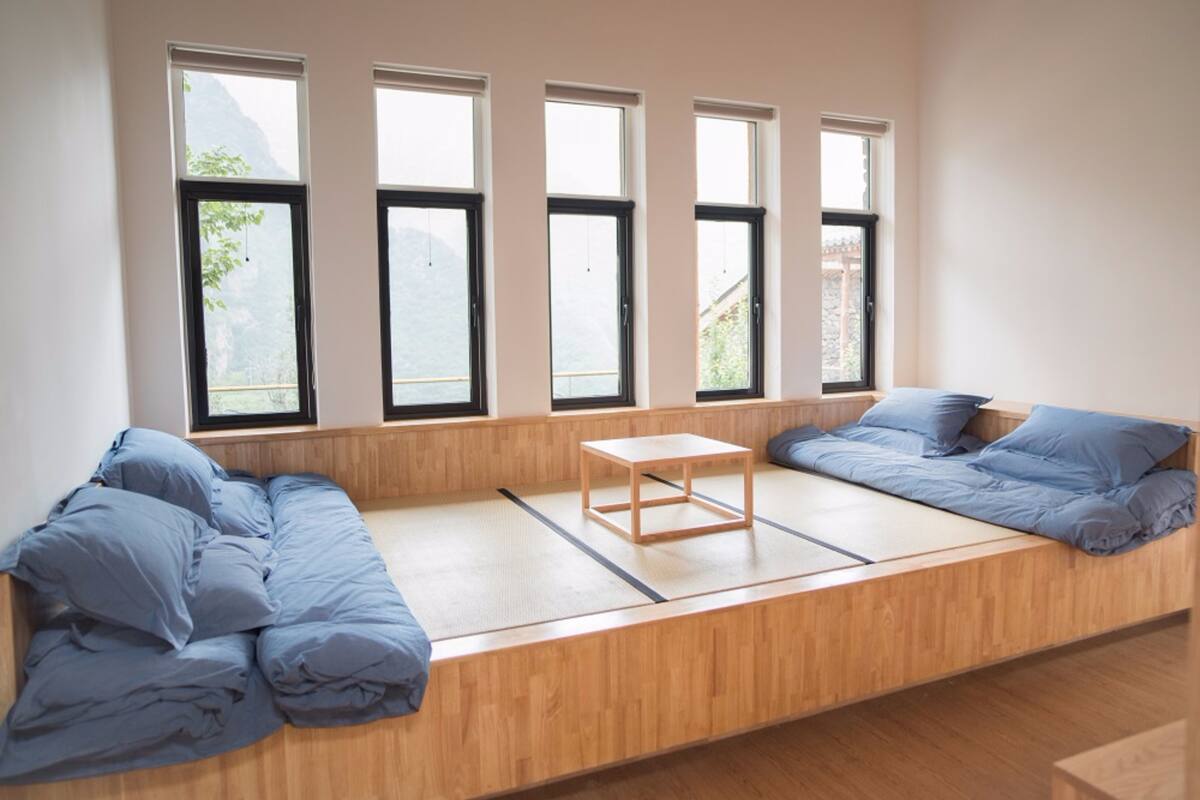 A room with a low wooden bed, a small wooden table, and large windows with a view of mountains.