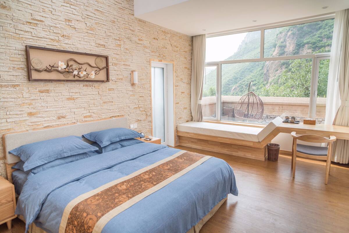 A bedroom with a large bed, a desk, and a window with a view of a mountainous landscape.