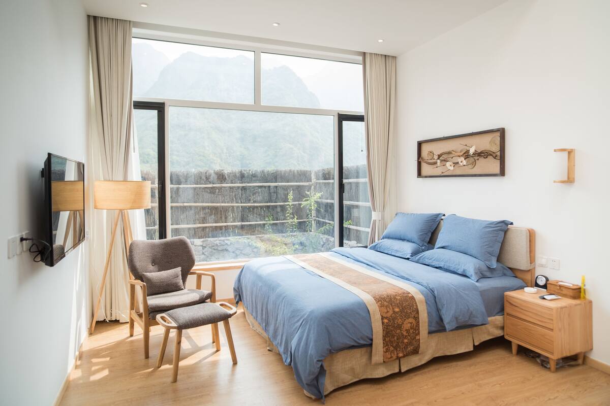 A bedroom with a large window, a bed, a chair, a bedside table, and a wall-mounted TV.