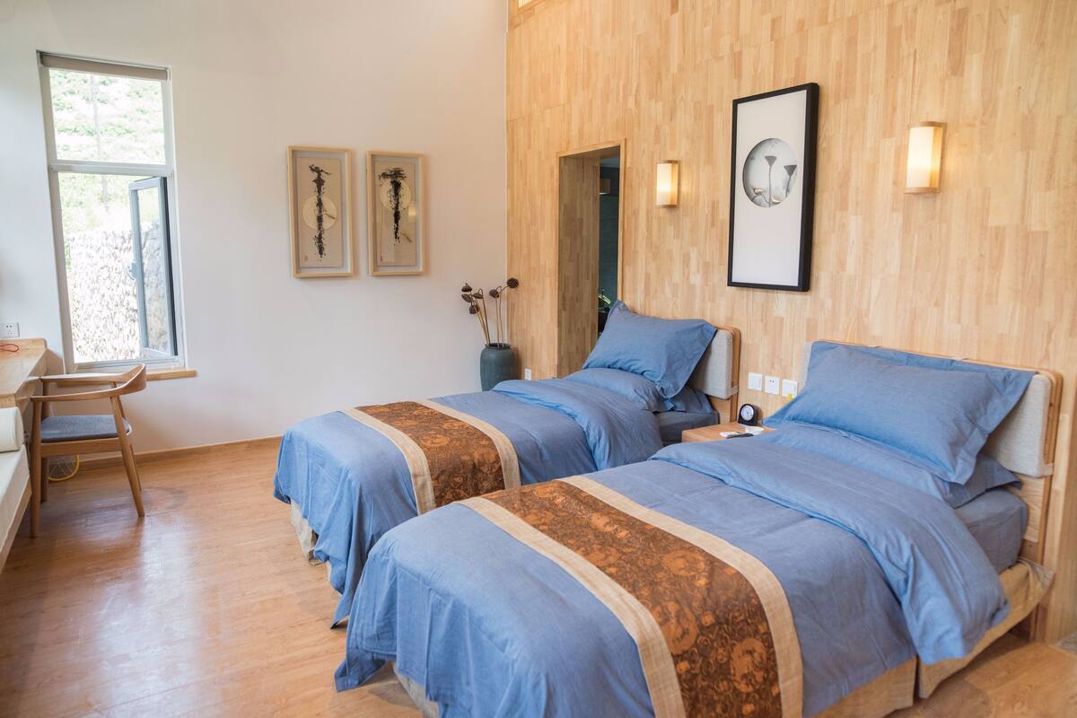 A bedroom with two beds, wooden flooring, a desk, and a chair. There are framed pictures on the wall and a vase with dried flowers.