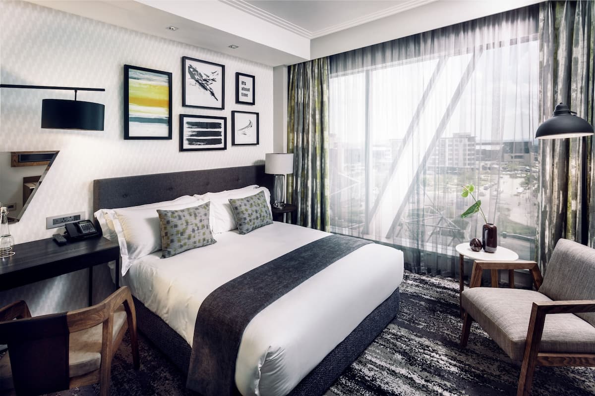 A hotel room with a large bed, a desk with a phone, a chair, a lamp, and a view of the city through sheer curtains.