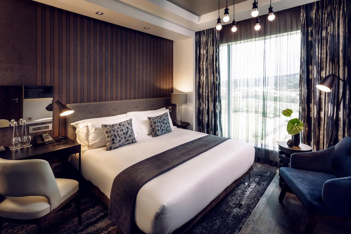 A modern hotel room with a large bed, a desk with a telephone, a chair, and a view of the outdoors through sheer curtains.