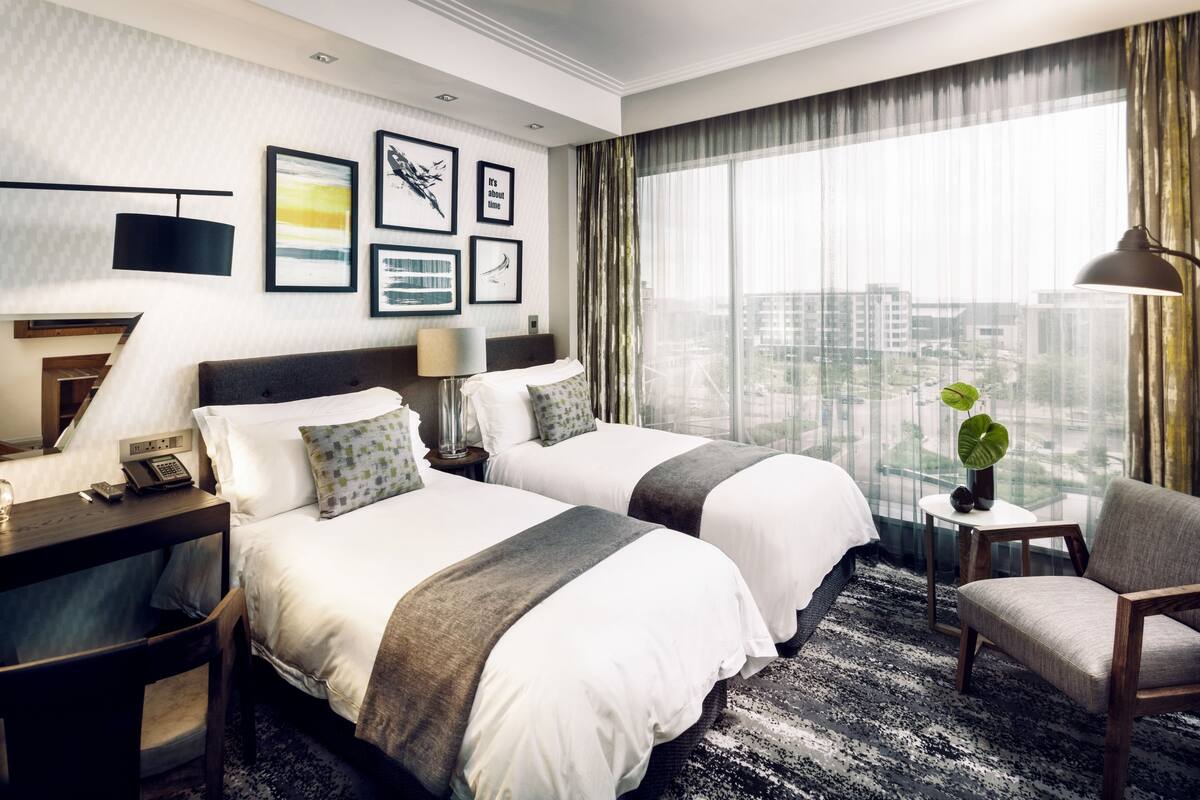 A hotel room with two beds, a desk, a chair, a lamp, and a view of the city.