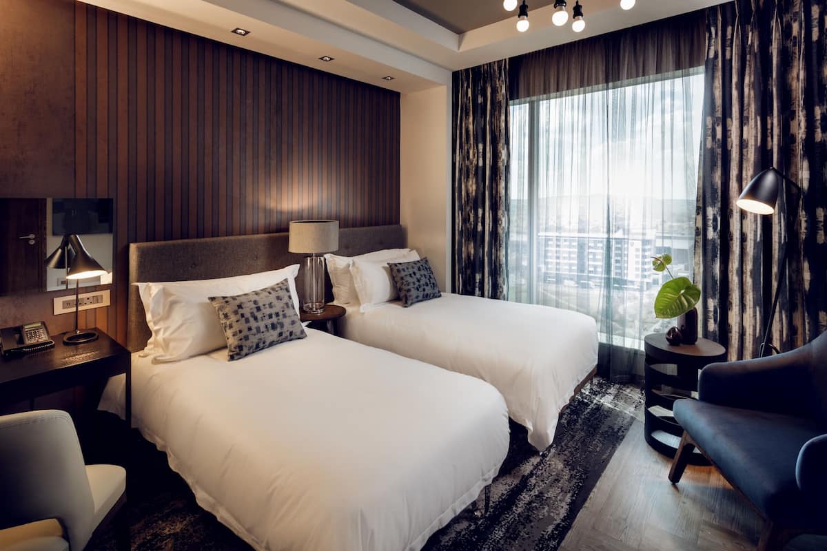 A hotel room with two beds, a desk with a lamp, a chair, a sofa, and a large window with curtains.