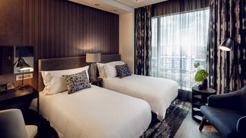 A hotel room with two beds, a desk with a lamp, a chair, a sofa, and a large window with curtains.