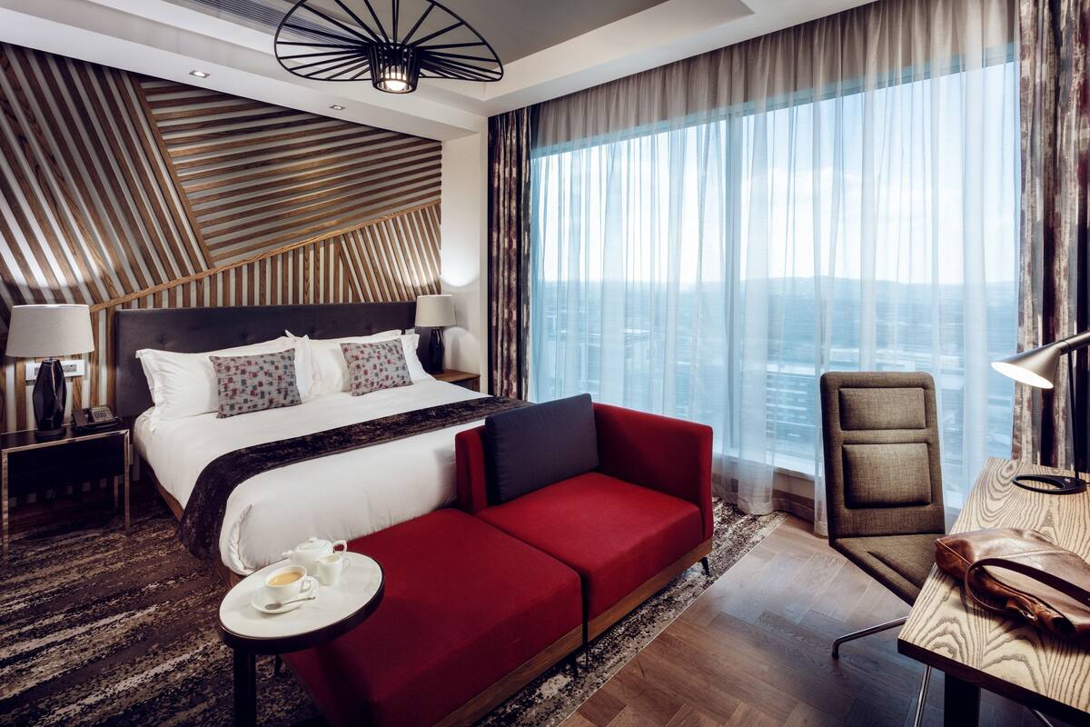 A modern hotel room with a large bed, a red sofa, a desk, and a view of the city.