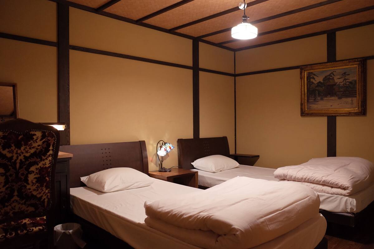 Habitación de hotel con dos camas individuales, una mesita de noche con lámpara, una silla y un cuadro en la pared.