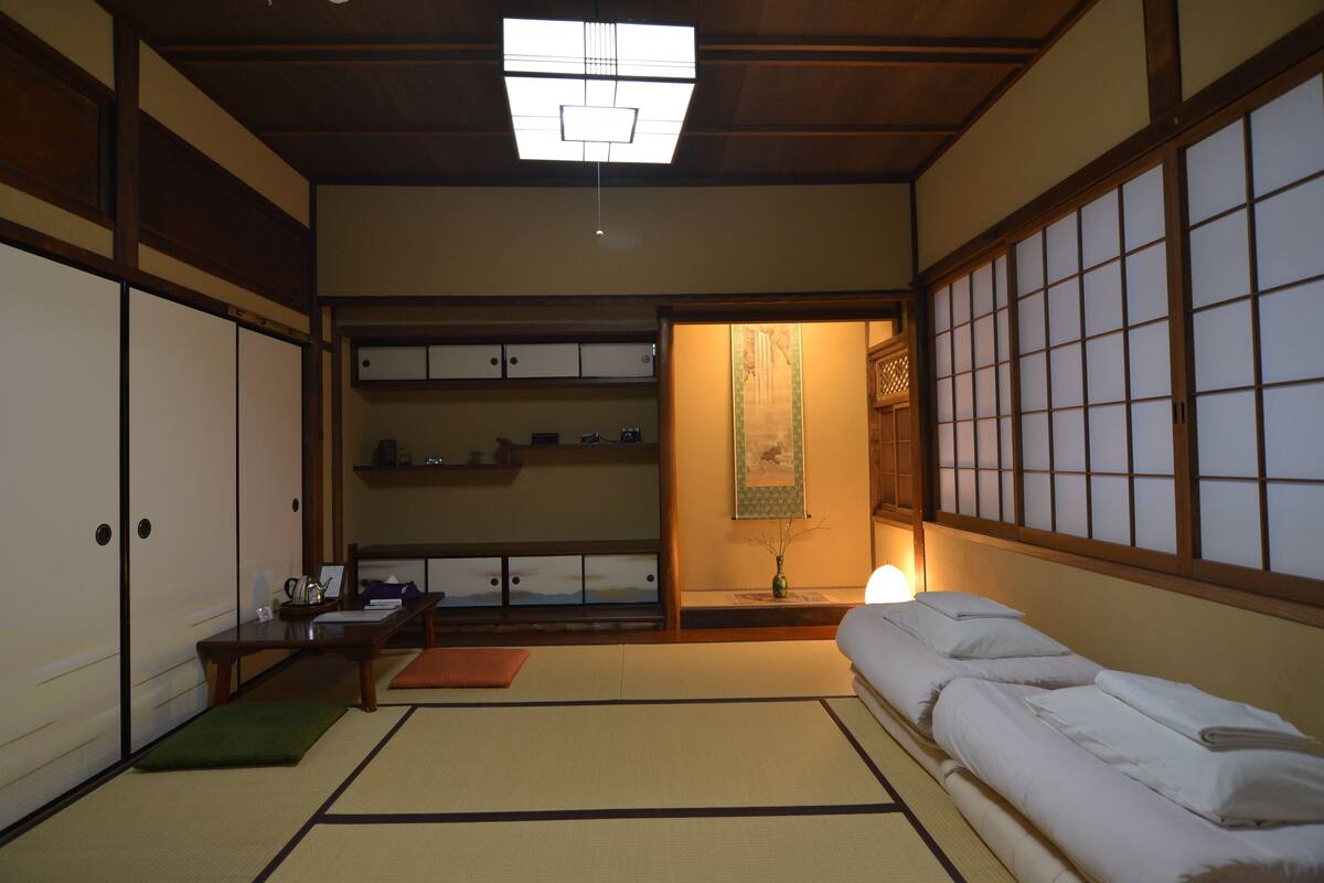 Una habitación japonesa tradicional con suelo de tatami, un futón, puertas correderas y una mesita baja con un jarrón y una lámpara.
