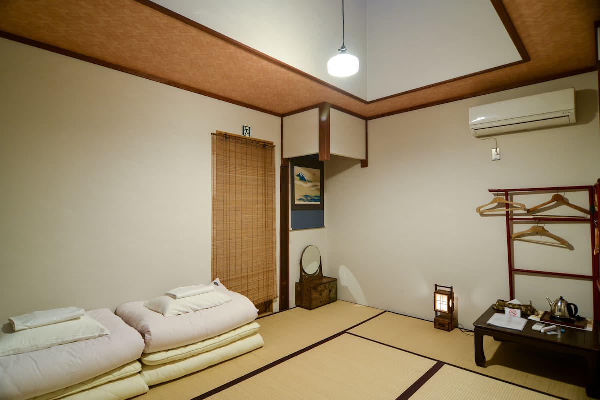 Una habitación japonesa tradicional con suelo de tatami, una cama futón, un estante de madera con perchas, una mesita con hervidor y un aire acondicionado.