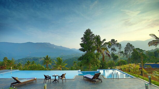 A pool area with lounge chairs and a table, surrounded by lush greenery and palm trees, overlooking a mountainous landscape. - The Leaf Munnar Resort (Devikolam)