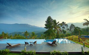A pool area with lounge chairs and a table, surrounded by lush greenery and palm trees, overlooking a mountainous landscape. - The Leaf Munnar Resort (Devikolam)
