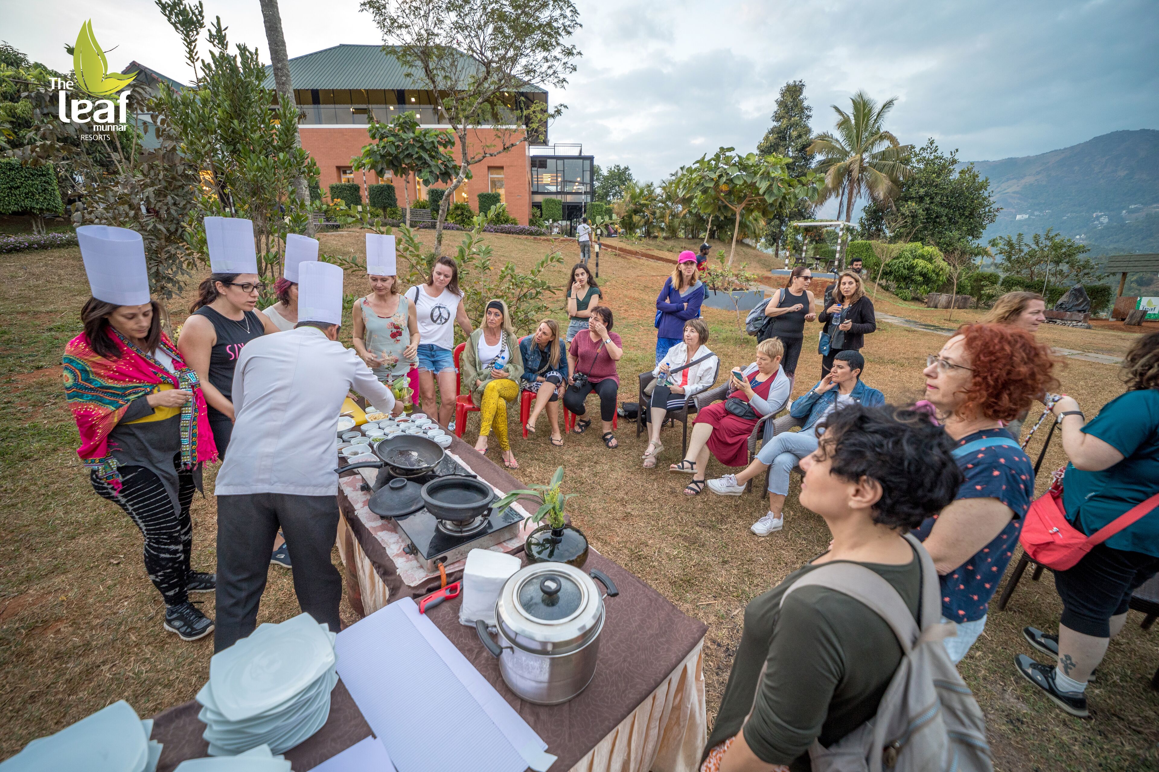 A group of people gathered around a cooking demonstration with a chef, outdoors with a view of a building and trees in the background.